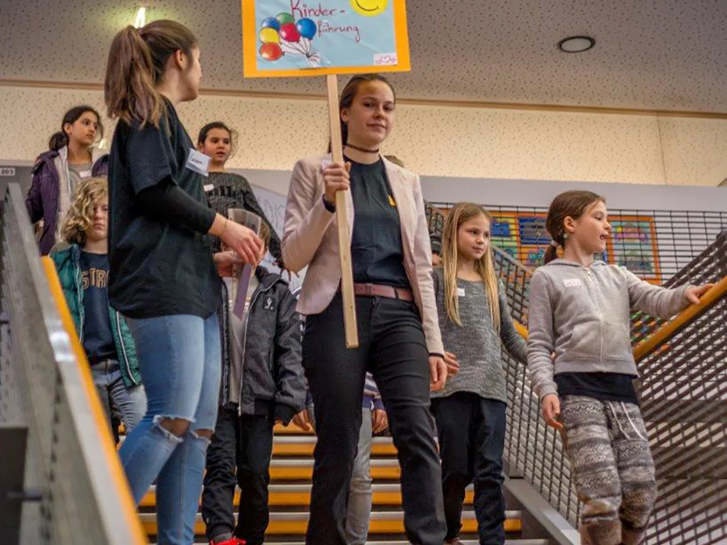 Kinder und eine Lehrkraft tragen ein Plakat die Treppe hinunter.